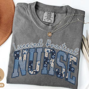 May include: A gray Comfort Colors t-shirt with the words "Licensed Practical Nurse" in white script and blue patchwork letters. A gold necklace with a pendant rests on the shirt. The shirt is size S-P-CH.
