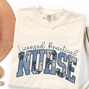 May include: Ivory-colored t-shirt with the text "Licensed Practical Nurse" in blue. The word "NURSE" is in a patchwork design with floral and gingham patterns. A gold necklace and a brown hat are visible.