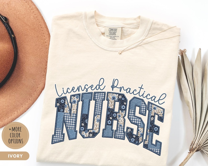 May include: Ivory-colored t-shirt with the text "Licensed Practical Nurse" in blue. The word "NURSE" is in a patchwork design with floral and gingham patterns. A gold necklace and a brown hat are visible.