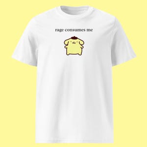 Puede incluir: Una camiseta blanca de manga corta con el gráfico de un pequeño perro de dibujos animados de color amarillo que lleva una boina marrón. Sobre el personaje, el texto "rage consumes me" está impreso en una fuente serif negra simple.