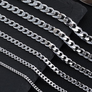 May include: Several silver curb chains of different widths are arranged on a black surface. The chains are displayed at an angle, showing their varying sizes and the reflective nature of the metal. The dark background accentuates the bright chains.