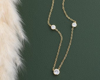 May include: A delicate gold necklace with six clear, round gemstones evenly spaced along the chain. The necklace is displayed on a dark green background, with a soft, feathery plant in the upper left corner.