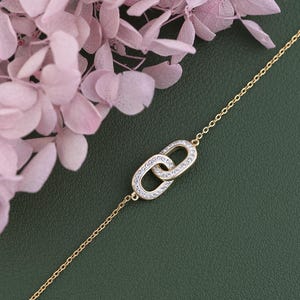 May include: A gold bracelet featuring two linked oval charms, each set with small, clear gemstones. The bracelet is displayed against a dark green backdrop, with pink flowers in the upper left. The chain is fine and delicate.