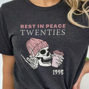 May include: Dark gray t-shirt featuring a graphic of a skull wearing a pink beanie and holding a coffee cup. The text "REST IN PEACE TWENTIES" and the year "1995" are printed above and below the graphic.
