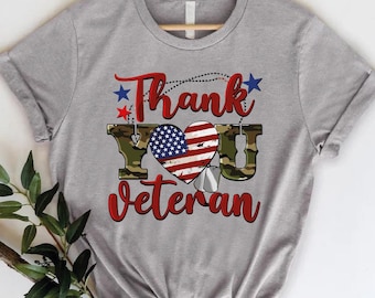 May include: A gray t-shirt with the words "Thank You Veteran" in red script. The design includes a heart with the American flag and camouflage lettering. Two dog tags and stars complete the patriotic theme.