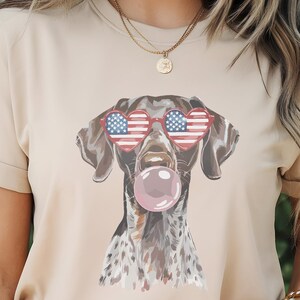 May include: A tan t-shirt featuring a graphic of a dog wearing heart-shaped sunglasses with an American flag design and blowing a pink bubble. The dog has brown and white spotted fur. The shirt has short sleeves.