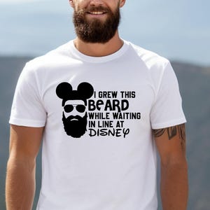 May include: White t-shirt with a black graphic of a beard with Mickey Mouse ears and sunglasses. The text reads, "I Grew This Beard While Waiting In Line At Disney."
