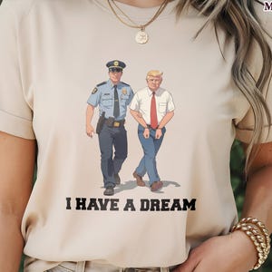 May include: Beige t-shirt featuring a cartoon illustration of a police officer and a man in a white shirt and red tie walking together. The text "I HAVE A DREAM" is printed below the image. The shirt has short sleeves.