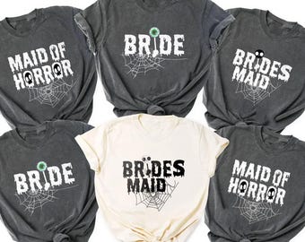 May include: A collection of gray and cream-colored t-shirts with Halloween-themed text. The shirts feature phrases like "Bride," "Brides Maid," and "Maid of Horror" in white, with spiderweb and skull graphics. The shirts are tied at the waist.