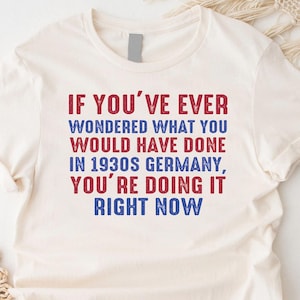 May include: A white t-shirt with a red and blue text graphic that reads "IF YOU'VE EVER WONDERED WHAT YOU WOULD HAVE DONE IN 1930S GERMANY, YOU'RE DOING IT RIGHT NOW".