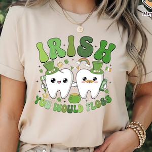 May include: Beige t-shirt with a St. Patrick's Day graphic. The design features two cartoon teeth wearing green top hats, with the text "IRISH YOU WOULD FLOSS" in green. Additional elements include a rainbow, shamrocks, and a pot of gold.
