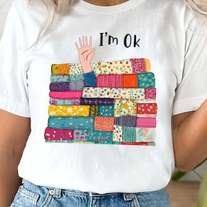 May include: White t-shirt with a colorful stack of books and a hand sticking out of the top. The text "I'm Ok" is printed on the shirt.
