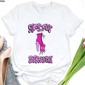 May include: White t-shirt featuring a pink high-heeled boot graphic with the text "STOMP OUT THE PATRIARCHY" in a stylized font. The shirt is displayed with a pair of light pink heels and a bouquet of pink tulips.