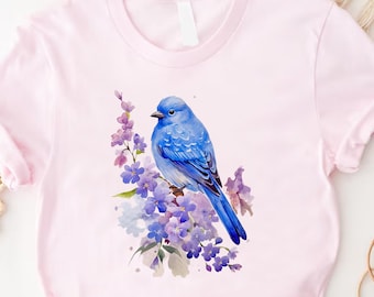May include: A light pink t-shirt featuring a watercolor illustration of a blue bird perched on a branch of purple flowers. The bird has detailed blue feathers and is set against a soft, pastel background.