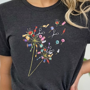 May include: A dark gray t-shirt with a graphic of a dandelion made of sewing supplies, including thread spools, needles, scissors, and buttons.