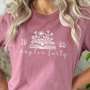 May include: A mauve t-shirt with a white graphic of an open book with flowers growing out of it. The text "19 chapter forty 85" is printed below the book.