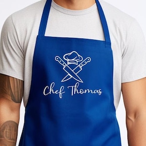 May include: A blue chef's apron with a white design of crossed chef's knives and a chef's hat with the text "Chef Thomas" underneath.