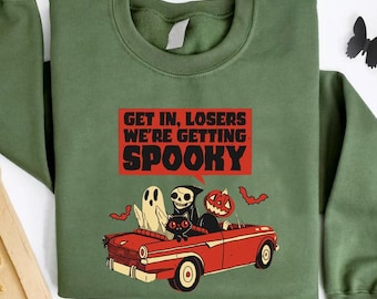 May include: Olive green sweatshirt with a Halloween-themed graphic. The graphic features a red car with a ghost, a skeleton, and a jack-o'-lantern. Text on the sweatshirt reads "GET IN, LOSERS WE'RE GETTING SPOOKY."