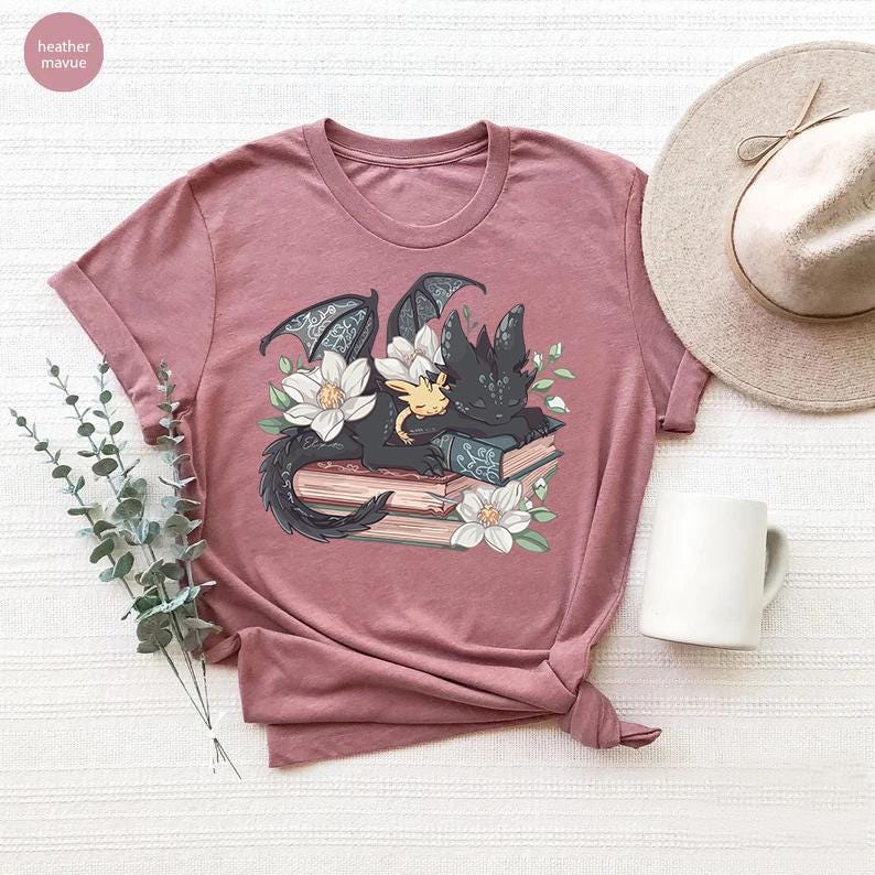 May include: A heather mauve t-shirt with a graphic of a black dragon and a yellow dragon sleeping on a stack of books with white flowers.