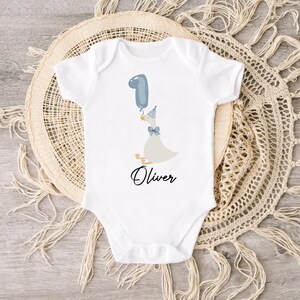 Puede incluir: Un body blanco para beb&eacute; con un globo azul, un pato con un gorro de fiesta azul y el nombre "Oliver" impreso en la parte delantera.
