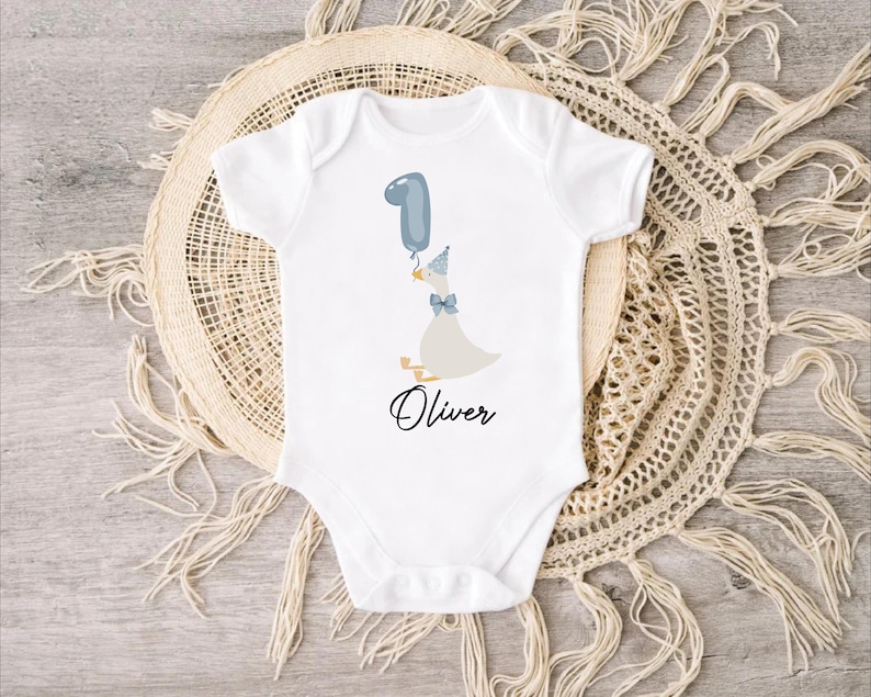 Puede incluir: Un body blanco para beb&eacute; con un globo azul, un pato con un gorro de fiesta azul y el nombre "Oliver" impreso en la parte delantera.
