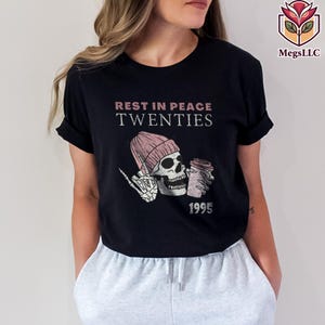 May include: Black t-shirt with the text "REST IN PEACE TWENTIES" above a graphic of a skeleton wearing a pink beanie and holding a coffee cup. The year "1995" is printed below the graphic. The shirt is a casual style.