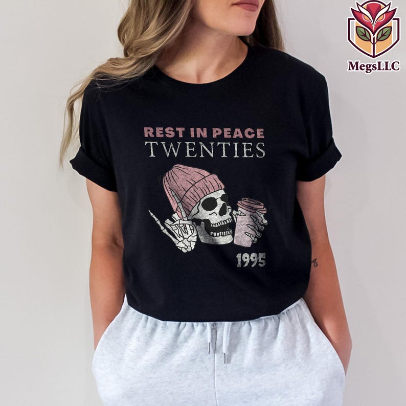 May include: Black t-shirt with the text "REST IN PEACE TWENTIES" above a graphic of a skeleton wearing a pink beanie and holding a coffee cup. The year "1995" is printed below the graphic. The shirt is a casual style.