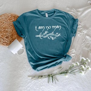 Puede incluir: Camiseta de color turquesa con la frase blanca "I am no man" y un dise&ntilde;o de espada y hojas. La camiseta est&aacute; anudada en la parte inferior. En el fondo se ven una taza blanca y un salvamanteles tejido.
