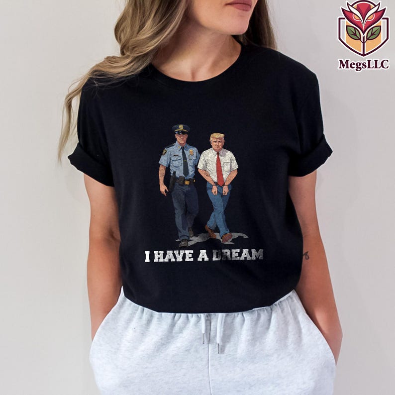 May include: Black t-shirt featuring an illustration of a police officer and a man in a white shirt and red tie. The text "I HAVE A DREAM" is printed in silver below the illustration. The shirt has short sleeves.