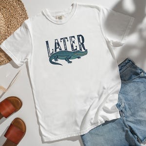 May include: White t-shirt with the word "LATER" in a distressed font above a blue and green alligator illustration. The shirt is paired with denim shorts, sandals, and a straw hat.