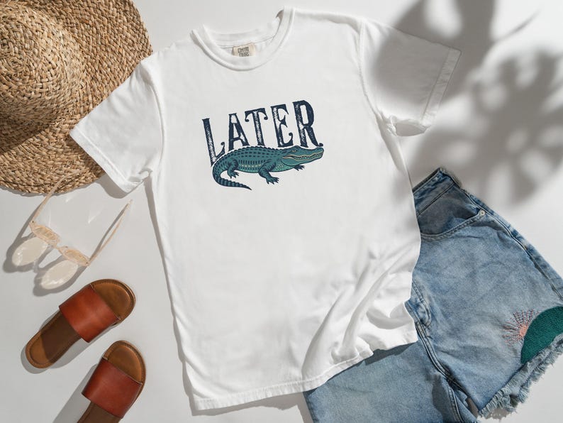 May include: White t-shirt with the word "LATER" in a distressed font above a blue and green alligator illustration. The shirt is paired with denim shorts, sandals, and a straw hat.