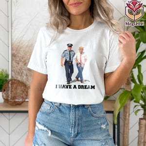 May include: White t-shirt featuring a graphic of a police officer and a man in a suit walking together. The text "I HAVE A DREAM" is printed in black below the image. The shirt is made of a soft, light-colored fabric.