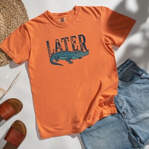 May include: Orange t-shirt with the word "LATER" above a blue alligator graphic. The shirt is paired with denim shorts, brown sandals, a straw hat, and sunglasses. A casual summer outfit.