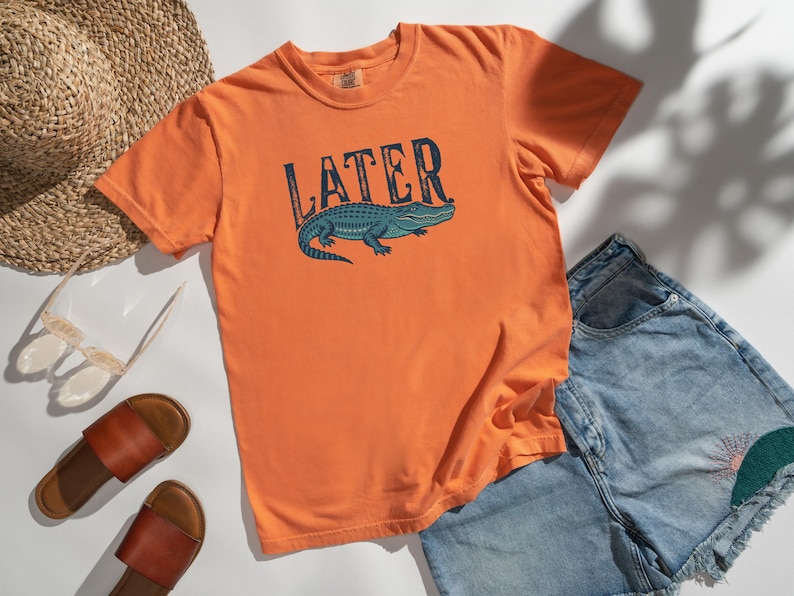 May include: Orange t-shirt with the word "LATER" above a blue alligator graphic. The shirt is paired with denim shorts, brown sandals, a straw hat, and sunglasses. A casual summer outfit.