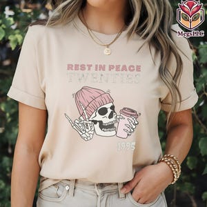 May include: Beige t-shirt featuring a graphic of a skull wearing a pink beanie and holding a coffee cup. The text "REST IN PEACE TWENTIES" and "1995" are printed above and below the skull illustration.