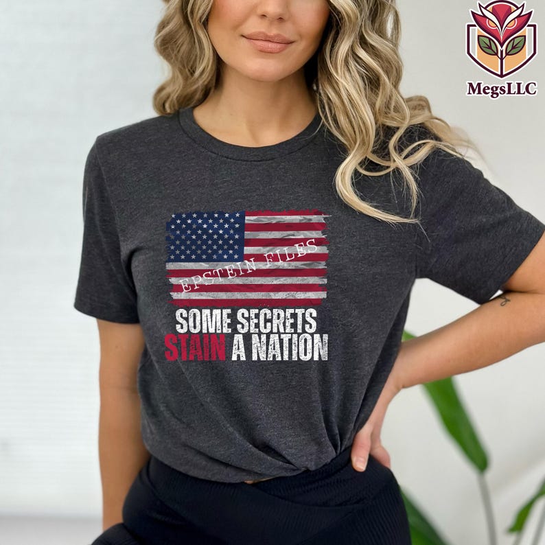 Pode incluir: T-shirt cinzenta escura com um design de bandeira americana desgastada e o texto "EPSTEIN FILES" e "SOME SECRETS STAIN A NATION" em branco e vermelho. A t-shirt tem gola redonda e mangas curtas.