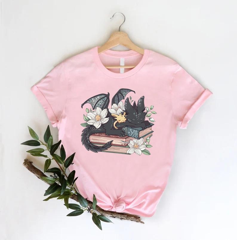 May include: A pink t-shirt with a black dragon and a small yellow dragon sleeping on a stack of books with flowers around them.