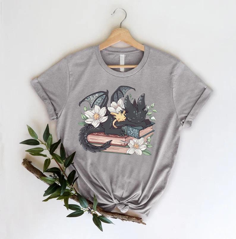 May include: A gray t-shirt with a black and yellow dragon sleeping on a stack of books with white flowers. The dragon has a small, yellow dragon curled up next to it.