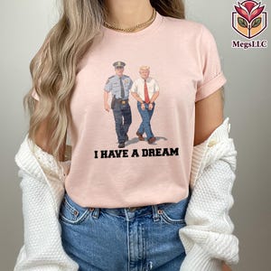 May include: A light pink t-shirt featuring an illustration of two men, one in a police uniform and the other in a suit, walking together. The text "I HAVE A DREAM" is printed in black below the illustration.