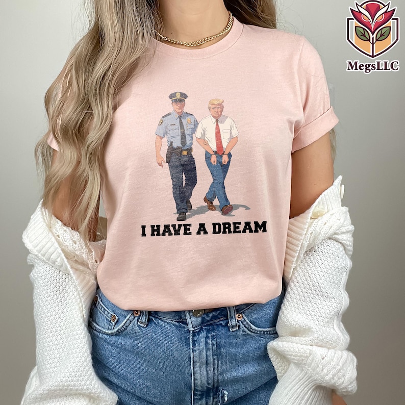May include: A light pink t-shirt featuring an illustration of two men, one in a police uniform and the other in a suit, walking together. The text "I HAVE A DREAM" is printed in black below the illustration.