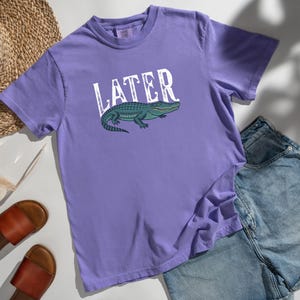 May include: A lilac-colored t-shirt with the word "LATER" in white block letters and a green alligator graphic. The shirt is paired with denim shorts, sandals, and a straw hat.