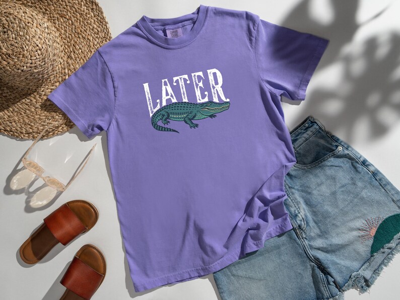 May include: A lilac-colored t-shirt with the word "LATER" in white block letters and a green alligator graphic. The shirt is paired with denim shorts, sandals, and a straw hat.