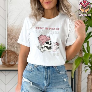May include: White t-shirt with a graphic design featuring a skull wearing a pink beanie and holding a coffee cup. The text "REST IN PEACE TWENTIES" and "1998" are printed above and below the skull.