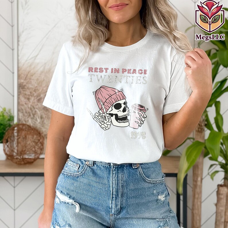 May include: White t-shirt with a graphic design featuring a skull wearing a pink beanie and holding a coffee cup. The text "REST IN PEACE TWENTIES" and "1998" are printed above and below the skull.