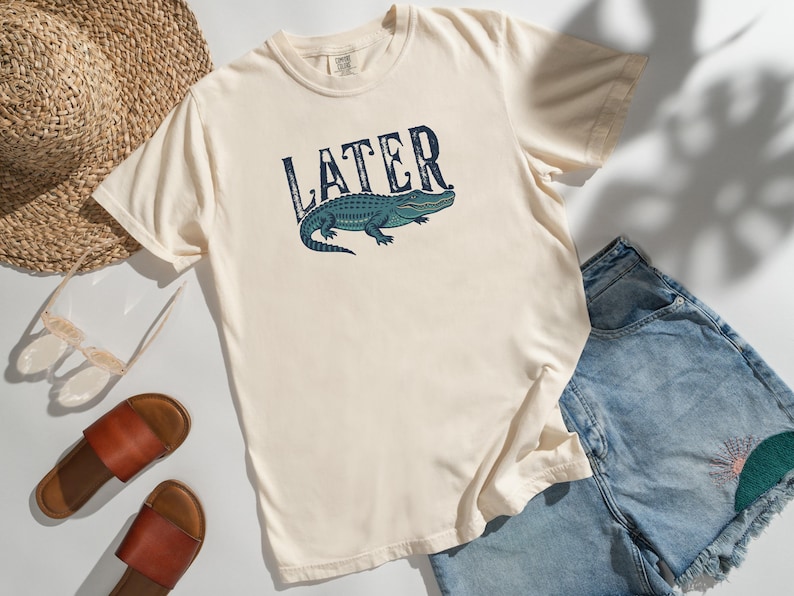 May include: Cream-colored t-shirt with the word "LATER" above a teal alligator graphic. The shirt is paired with denim shorts, brown sandals, a straw hat, and sunglasses, suggesting a casual, summery theme.