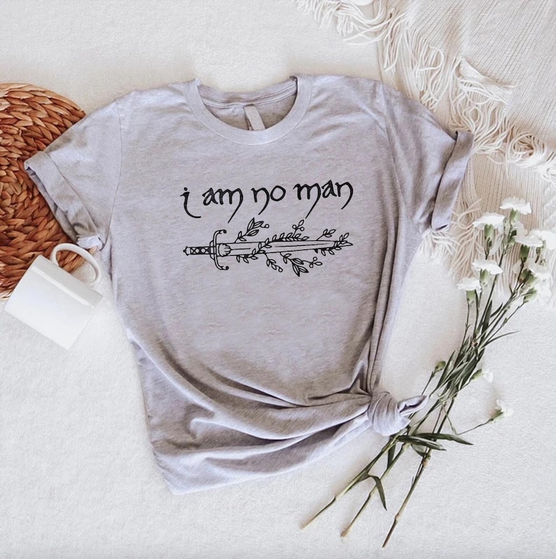 Puede incluir: Camiseta gris claro con el texto "I am no man" sobre un dise&ntilde;o de espada y hojas en negro. La camiseta est&aacute; anudada en la parte inferior derecha. Una taza blanca y flores en el fondo.