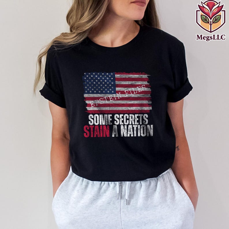 Pode incluir: Camiseta preta com um design de bandeira americana desgastada e o texto "EPSTEIN FILES". Abaixo, a frase "SOME SECRETS STAIN A NATION" &eacute; impressa em branco e vermelho. Gola redonda.