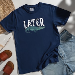 May include: Navy blue t-shirt with the word "LATER" in white, above a teal alligator illustration. The shirt is paired with denim shorts, brown sandals, a straw hat, and sunglasses.
