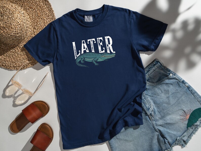 May include: Navy blue t-shirt with the word "LATER" in white, above a teal alligator illustration. The shirt is paired with denim shorts, brown sandals, a straw hat, and sunglasses.