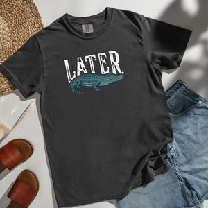 May include: Dark gray t-shirt with the word "LATER" in white, featuring a blue and green alligator illustration. The shirt is paired with denim shorts, sandals, and a straw hat.